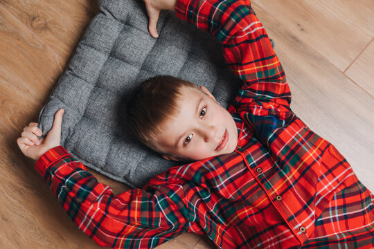 A cheerful boy in red checkered pajamas lies on a wooden floor on a gray pillow. View from above. Preparing for Christmas