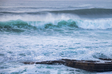 A powerful ocean wave crashes and breaks, creating foamy white spray as it approaches a rocky shoreline under an overcast sky. 