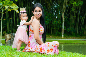 latina mother with her beautiful brunette baby, sitting by a lake next to a giant bamboo forest, woman looking proudly at her little daughter.