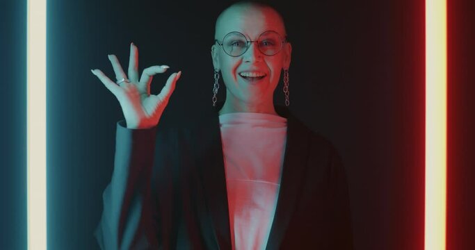 Slow motion portrait of satisfied lady making OK hand gesture and smiling on black background with neon illumination. People and positive emotion concept.