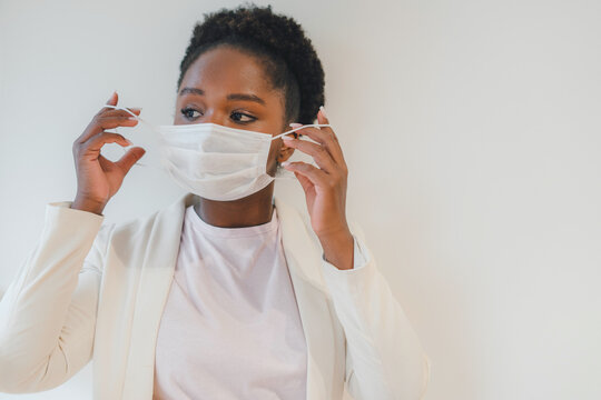 Portrait of african-american woman in white jacket putting on medical face mask and looking away isolated over with wall. Self isolation, prevention, quarantine