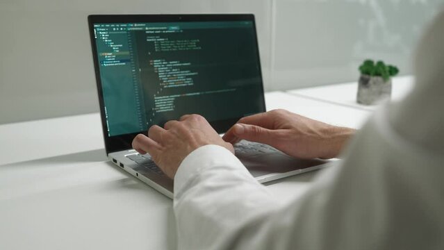 Over the shoulder view of professional developer coding in laptop working in IT company programming. Hands of man IT worker typing on keyboard doing tasks developing checking code.