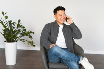 Cheerful middle aged asian man calling by phone, sitting in armchair in living room interior with white wall