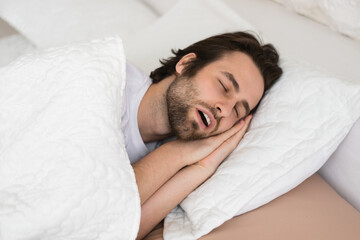 Tired young caucasian male with open mouth sleeping on white soft bed in bedroom in spare time, enjoy quiet
