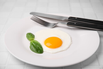 Tasty fried egg with basil in plate on white tiled table, closeup