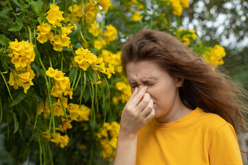 Portrait of beautiful young allergic woman is suffering from pollen allergy or cold on natural flower flowering tree background at spring or sunny summer day sneezes, blowing her runny nose rubs eyes - obrazy, fototapety, plakaty