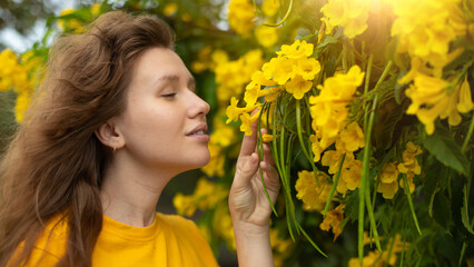 Portrait of happy beautiful bearded girl, young positive woman with beard is smelling beautiful yellow flowers in the garden, smiling, enjoying spring or summer day, breathing deep deeply fresh air