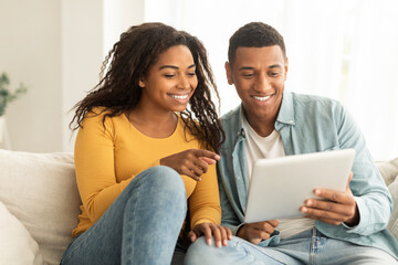 Cheerful millennial african american wife show finger at husband tablet, have video call in living room interior