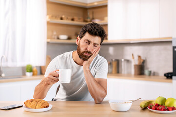 Tired sad sleepy handsome millennial caucasian guy with beard in domestic clothes with cup of coffee sleeps at table
