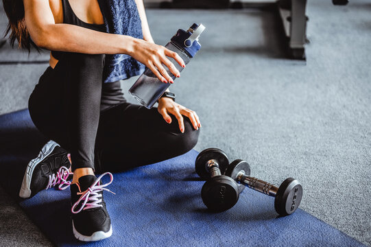 Fitness asian woman relaxing after exercise with drinking waters and dumbbell placed beside the gym.Relaxing after training.beautiful young woman looking away while sitting at Fitness gym.Fitness
