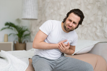 Despaired sad young caucasian guy suffers from chest pain, heart attack, sits on white bed alone in bedroom