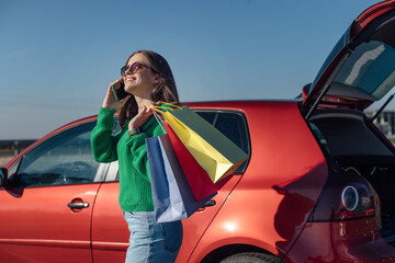 Woman in green sweater talking on smartphone and holding on shoulders paper bags near car's trunk