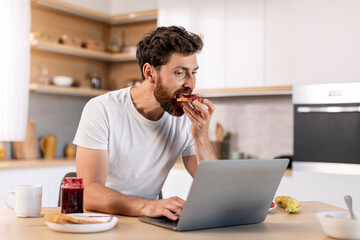 Fototapeta na wymiar Busy millennial caucasian man with beard eats sandwich with jam and works on computer, surfing in internet