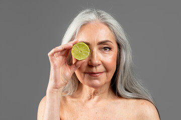 Serious old european female with gray hair puts lemon to eye, isolated on gray background, studio