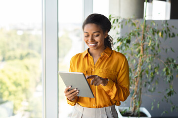 Happy black female entrepreneur using tablet standing near window at modern coworking office, copy...