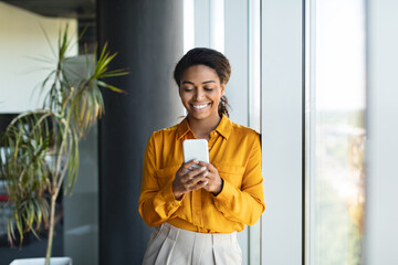 Happy successful black businesswoman typing on smartphone, standing near window in modern office...