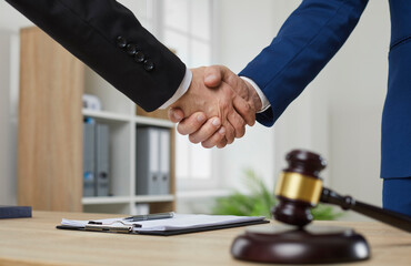 Professional lawyer shaking hands with client. Two men in suits exchange handshakes over office table with judge's gavel. Closeup shot of human hands. Law consultation, legal services concept