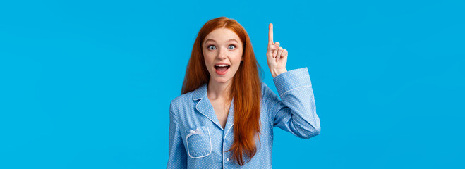 Fototapeta na wymiar I got solution, hear me out. Attractive excited redhead girl in nightwear sharing her ideas, raising index finger eureka gesture, talking to you, saying plan, standing in pyjamas, blue background