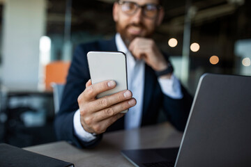 Middle aged businessman sitting at desk and holding cellphone in hand, texting with clients, selective focus