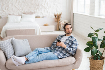 Glad pensive young caucasian guy lying on sofa with smartphone, chatting in social networks, watch...