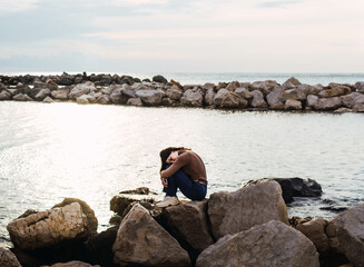 woman sitting on stones in front of the sea hugging her legs at sunset