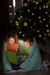 children under a beautiful Christmas tree read a book. a boy and a girl play under a Christmas tree decorated with oranges