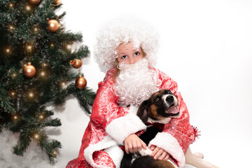 a cute child in a New Year's hat hugs a kind dog near the New Year's tree. New Year's bag with gifts lies under the Christmas tree. girl dressed as santa claus hugging a beautiful dog