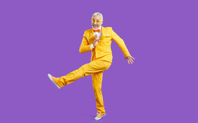 Fototapeta na wymiar Overjoyed mature active man in yellow suit isolated on purple studio background dancing. Happy energetic old pensioner in jacket make dancer moves. Elderly party and celebration.