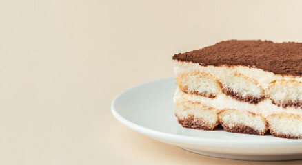 Homemade tiramisu cake on white plate against beige background. Traditional Italian no-bake dessert made of savoiardi, filled with mascarpone cheese, coffee espresso and sprinkled with cocoa powder