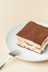 Homemade tiramisu cake on white plate against beige background. Traditional Italian no-bake dessert made of savoiardi, filled with mascarpone cheese, coffee espresso and sprinkled with cocoa powder