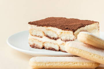 Homemade tiramisu cake on white plate against beige background. Traditional Italian no-bake dessert made of savoiardi, filled with mascarpone cheese, coffee espresso and sprinkled with cocoa powder