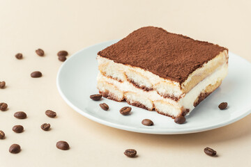 Homemade tiramisu cake on white plate against beige background surrounded by coffee beans . Italian dessert made of savoiardi cookies filled with mascarpone cheese and sprinkled with cocoa powder