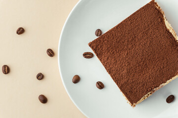 Tiramisu cake on white plate against beige background surrounded by coffee beans . Italian dessert made of savoiardi cookies filled with mascarpone cheese and sprinkled with cocoa powder. Flat lay