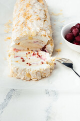 Cut of homemade meringue roll decorated with almond flakes and freeze-dried red berries on the white marble background. Meringue roulade with whipped cream on a parchment paper