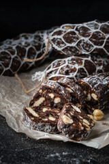Chocolate salami filled with almonds, hazelnuts and raisins on the black background. Italian sweet sausage shaped dessert sprinkled with powdered sugar. Christmas and New Year festive pastry