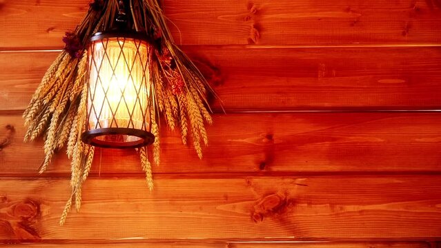 Beautiful warm cozy background warming the soul. Glowing lantern on the wall of a wooden house. Village hut. A bouquet of dry ears and flowers is hung on the wall from wooden boards. Heat-emitting