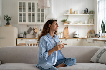 Carefree young woman holding cup of coffee sits on sofa at home, dreaming, looking into distance enjoying loneliness. Optimistic Italian female dressed in casual style contemplates future of weekend
