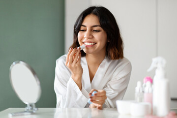 Cheerful cute young woman using lipbalm, applying makeup at home