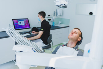 A young man is sitting in a dental chair, and a dentist is working at a computer. Dentist's appointment