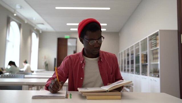 Talented young black guy scientist wearing glasses studying in library, reading book and taking notes. Focused African American male student writing research summary, sitting at desk. Education