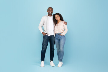 Portrait of happy black adult couple embracing, looking at camera and smiling, posing over blue...