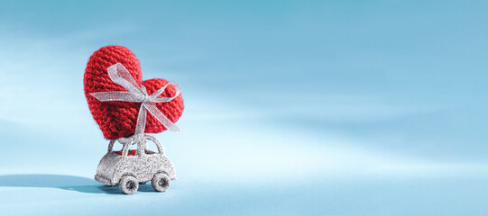 Glittery toy car delivering red knitted craft heart on roof top on blue background. St Valentines Day thank you card Mothers or Fathers Day. Birthday gift or sales delivery banner
