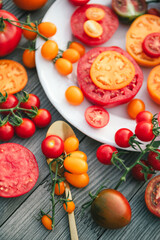 Delicious raw fresh colorful tomatoes of different shapes on white round plate with spoon on wooden background. Chopped red and yellow tomatoes. Healthy food rich in vitamin C