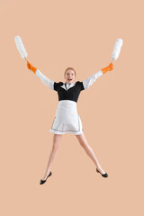 Young female chambermaid with dusters jumping on beige background