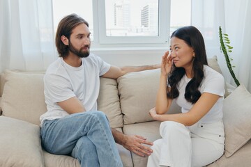 A man and a woman of different races sit on the couch in a room at home and talk about their problems to each other. A stress-free lifestyle of family quarrels with psychological support