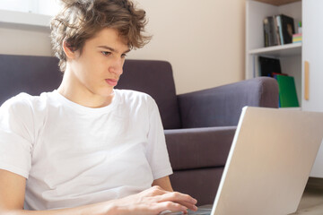 Cute smiling young teenager with laptop on the couch at home. Boy in white t shirt makes homework with a computer. Distance, online education for children