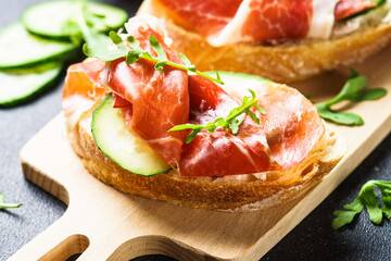 Appetizer crostini, open sandwiches with cream cheese, prosciutto, cucumber and arugula at wooden table. Flat lay.