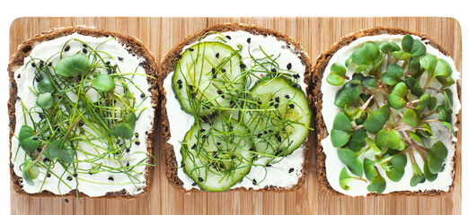 Healthy breakfast with micro greens. Close-up top view of toasts on brown bread with home grown microgreens, cucumber and fresh cream cheese on wooden board.