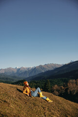 Fototapeta premium Woman full-length sitting resting on a hill and looking at the mountains in a yellow raincoat and jeans happy camping trip in the winter, freedom lifestyle 