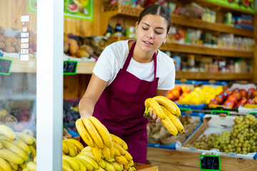 Positive young European woman in apron picking out ripe bananas from wooden box in hypermarket